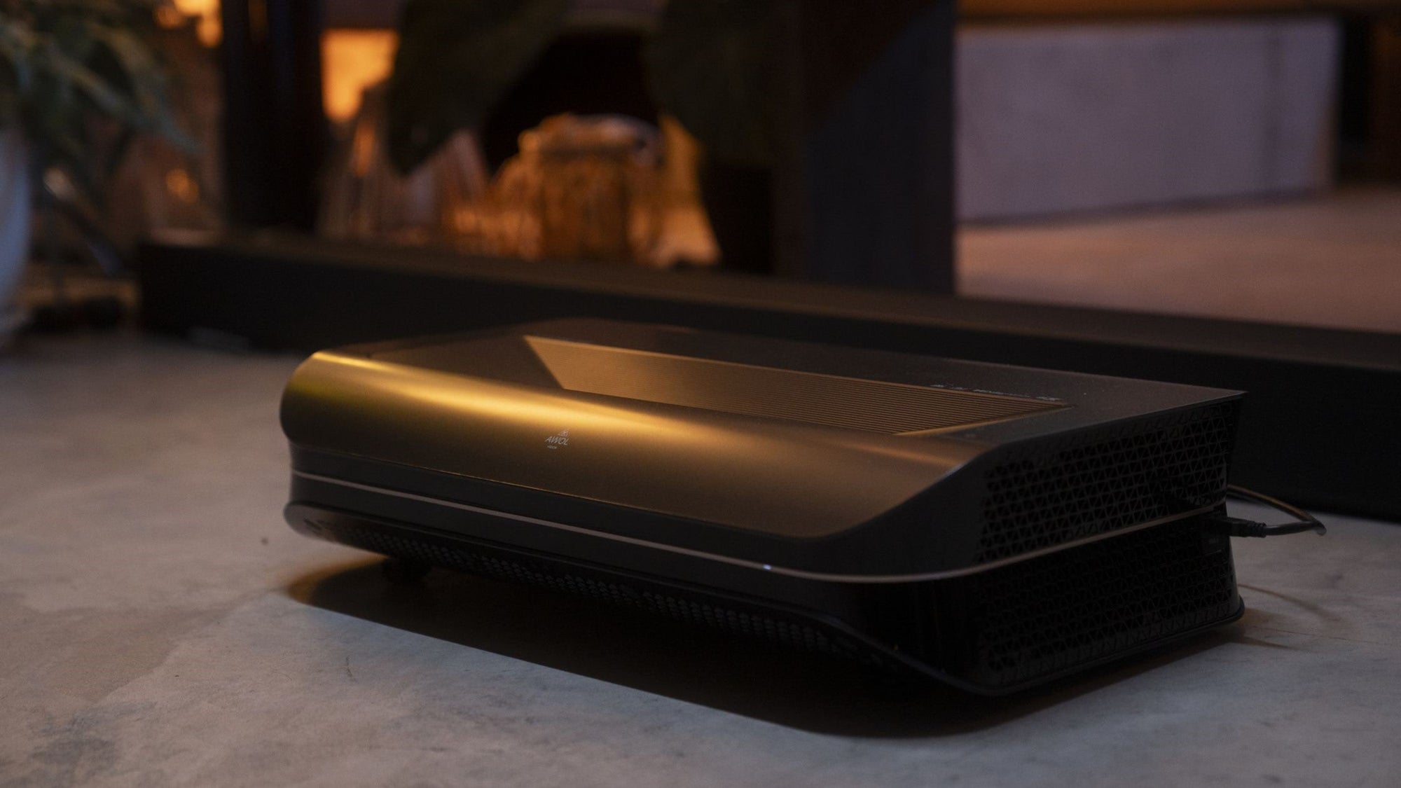 A sleek, dark ultra-short throw (UST) projector sits on a light-colored floor in a modern living room setting, casting an unseen image upwards.